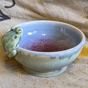 May include: A small ceramic bowl featuring a sculpted green frog perched on the rim. The interior of the bowl has a light blue glaze that transitions into a deep purple center, while the exterior is finished in a muted sage green glaze.