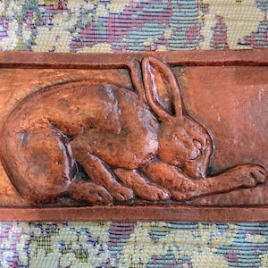 May include: A rectangular terracotta-colored wall plaque featuring a relief sculpture of a resting rabbit. The rabbit is depicted in a curled position with its ears upright and head tucked toward its front paws, set within a simple raised border frame.