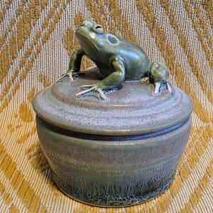 May include: A round ceramic lidded jar with a glossy green glaze. The lid features a sculpted, three-dimensional green frog perched on top. The jar has a smooth, tapered body with a mottled finish that transitions from light to dark green.