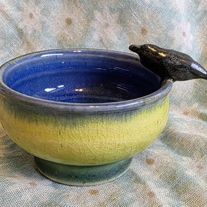 May include: A ceramic bowl with a blue interior and a yellow and green exterior. A dark gray seal figurine is perched on the rim. The bowl has a pedestal base and a crackled glaze finish, adding a unique texture to the piece.