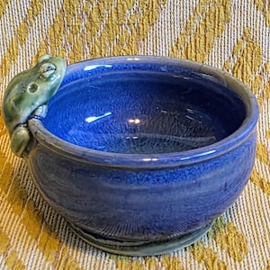May include: A round ceramic bowl with a glossy, deep blue glaze featuring a sculpted, light green frog perched on the outer rim. The bowl has a smooth, rounded interior and a slightly flared edge, showcasing a vibrant, reflective finish throughout.