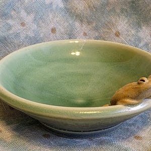 May include: A round ceramic bowl with a smooth, glossy light green glaze. A small, sculpted tan frog figure is attached to the inner rim, appearing to peer over the edge into the center of the bowl. The bowl has a wide, shallow basin shape.