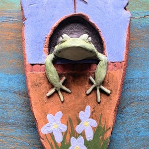 May include: A hand-painted clay wall plaque shaped like a shield, featuring a light green frog emerging from a dark arched opening. The top section is light blue with a carved rune, while the bottom is terracotta brown with green grass and three light purple flowers.