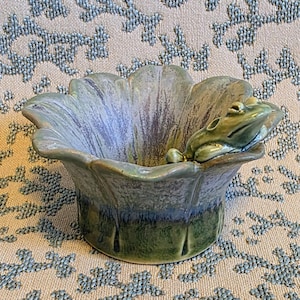 May include: A ceramic bowl shaped like a flower with scalloped edges, featuring a glazed finish in shades of sage green, lavender, and deep blue. A small, sculpted green frog is perched on the rim of the bowl, facing inward.