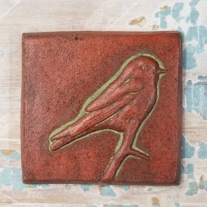 May include: A red ceramic tile with a green outline of a bird perched on a branch. The bird is facing the right side of the tile.