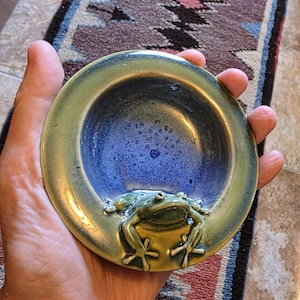 May include: A small, round ceramic bowl featuring a glossy, deep blue interior and a wide, olive-green rim. A sculpted, three-dimensional green frog is attached to the inner edge of the bowl, with its front legs resting on the rim and its head facing inward.