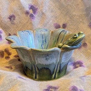May include: A ceramic bowl shaped like a flower with scalloped edges, featuring a glazed finish in shades of blue, green, and white. A small, sculpted green frog is attached to the rim of the bowl, resting its head on the edge of the petal.