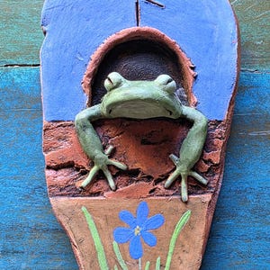 May include: A teardrop-shaped clay plaque featuring a sculpted green frog emerging from a dark arched opening. The top section is painted blue with a carved rune symbol. The bottom section is brown with a painted blue flower and green stems.