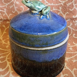 May include: A cylindrical ceramic jar with a lid featuring a sculpted green frog figurine on top. The jar has a glossy glaze that transitions from a deep black at the base to a vibrant blue in the middle and a lighter blue on the lid.