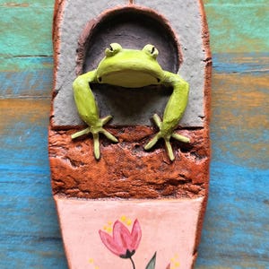 May include: An elongated, oval-shaped clay plaque featuring a sculpted green frog emerging from a gray arched opening. Above the arch is an etched F-shaped rune. The lower section is painted light pink with a hand-painted pink flower and green leaves.