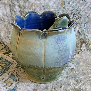 May include: A ceramic planter with a scalloped rim and a sculpted frog perched on the edge. The exterior features a gradient of sage green and earthy brown tones, while the interior is finished in a deep, glossy cobalt blue glaze.