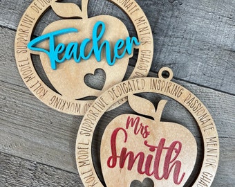 Può includere: Due ornamenti in legno a forma di mela con le parole "Teacher" e "Mrs. Smith" scritte in blu e rosso, rispettivamente. Gli ornamenti sono decorati con un cuore e circondati da un cerchio di testo che descrive le qualità di un insegnante.