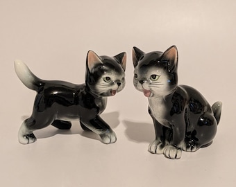 May include: Two ceramic cat figurines featuring a glossy black and white coat. One cat is depicted in a walking stance with its tail raised, while the other is sitting upright. Both cats have painted green eyes, pink ears, and open mouths.
