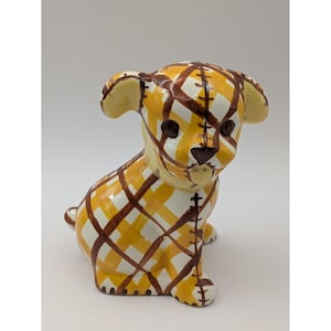 May include: A ceramic puppy figurine in a sitting position. The puppy is painted with a yellow, white, and brown plaid pattern. The ears and nose are a solid color. The figurine has a stitched design on the sides.