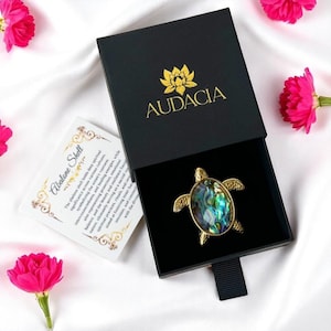 Puede incluir: Un colgante dorado en forma de tortuga con una incrustación de concha de abulón, presentado en una caja negra abierta con el nombre de la marca "AUDACIA" en letras doradas. También es visible una tarjeta con texto sobre las conchas de abulón.