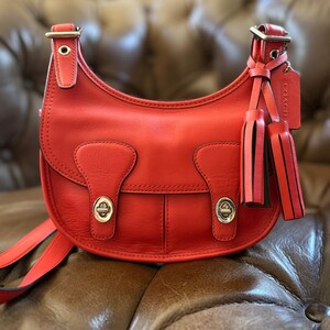 May include: A bright coral red leather crossbody bag featuring a curved silhouette, two front flap pockets with silver-tone turn-lock closures, an adjustable shoulder strap, and a matching leather tassel charm with an embossed Coach brand tag.