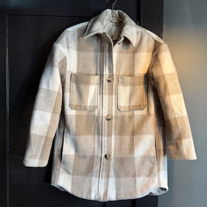May include: A beige and white plaid jacket with a collared neckline, two chest pockets, and button closures. The jacket is hanging on a gold hanger against a dark background. The fabric appears to be a soft, woven material.