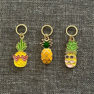 Peut inclure: Trois breloques ananas dorées avec des motifs différents. Un breloque ananas a des lunettes de soleil en forme de cœur rouge, un autre a une couronne verte et des lunettes de soleil, et le dernier a un design d'ananas simple.