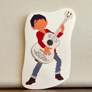 K&ouml;nnte beinhalten: Ein Aufkleber mit einer Cartoon-Illustration eines Jungen, der eine wei&szlig;e Gitarre spielt. Der Junge tr&auml;gt ein rotes Hemd, blaue Hosen und braune Schuhe. Die Gitarre hat ein dekoratives Design.