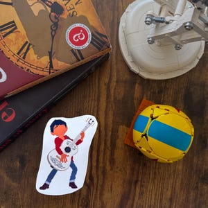 K&ouml;nnte beinhalten: Ein Aufkleber mit einer Cartoonfigur, die eine wei&szlig;e Gitarre spielt, blaue Jeans und ein rotes Hemd tr&auml;gt. Ein gelb-blauer geometrischer Ball und ein Buch mit dem Titel "The Seven Dials Mystery" sind ebenfalls zu sehen.