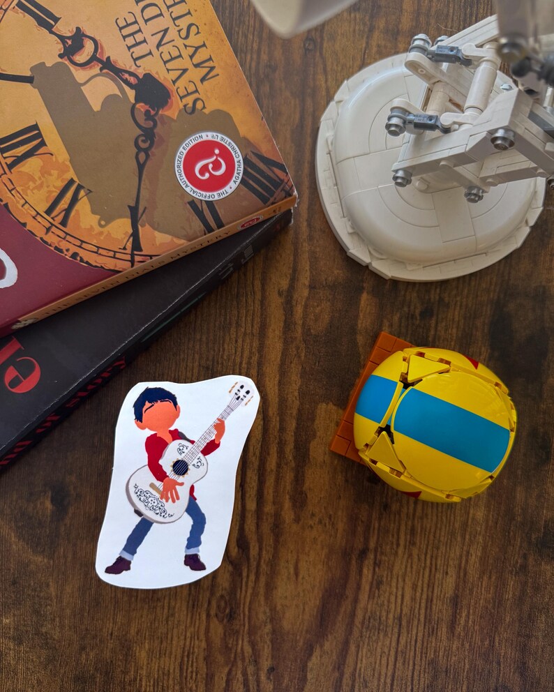 K&ouml;nnte beinhalten: Ein Aufkleber mit einer Cartoonfigur, die eine wei&szlig;e Gitarre spielt, blaue Jeans und ein rotes Hemd tr&auml;gt. Ein gelb-blauer geometrischer Ball und ein Buch mit dem Titel "The Seven Dials Mystery" sind ebenfalls zu sehen.