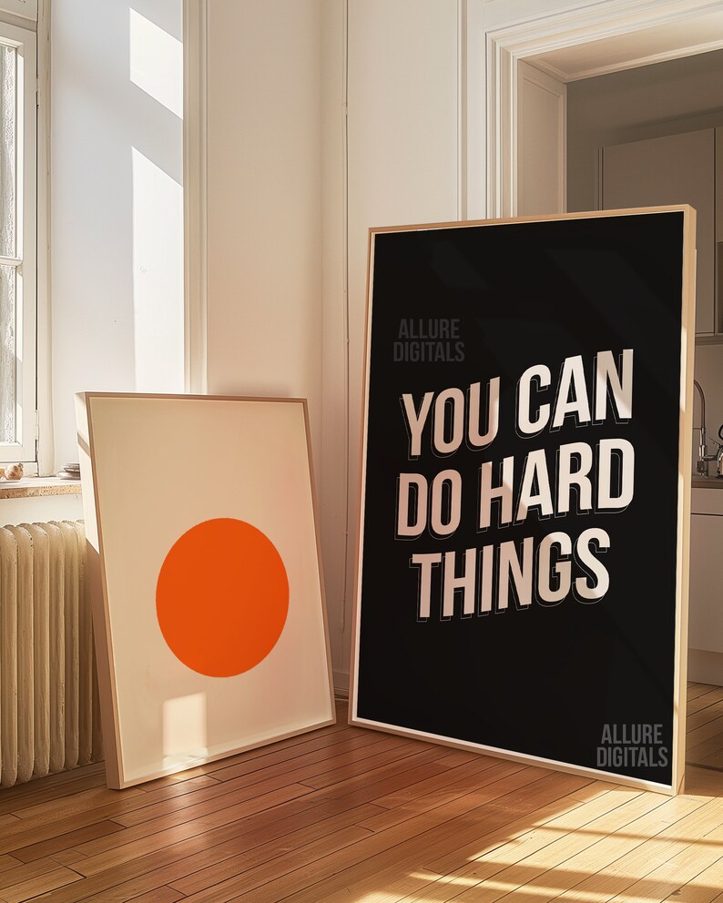May include: Two framed prints on a wooden floor. The print on the left is a simple white background with a large orange circle in the center. The print on the right is black with white text that reads "You can do hard things." The text is in a bold, sans-serif font. The text "Allure Digitals" appears at the top and bottom of the print.