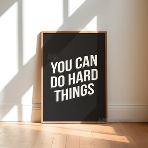 May include: A black framed poster with white text that reads "You can do hard things". The poster is on a wooden floor with a white wall in the background.