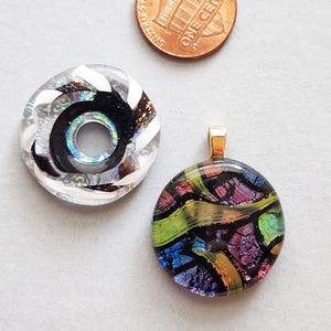 May include: Two dichroic glass pendants. One is a clear circle with a black spiral and white accents. The other is a round pendant with a gold bail, featuring a colorful abstract design with green, purple, and blue hues. A penny is in the background.