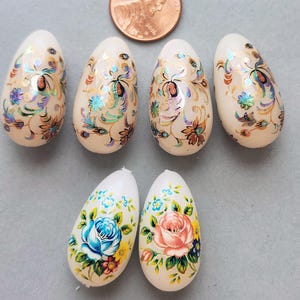 May include: Six decorative egg-shaped ornaments. The top row features eggs with gold and iridescent floral designs. The bottom row displays eggs with blue and pink rose patterns. A penny is included for size reference.