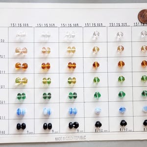May include: A display card showcasing a variety of colorful glass beads. The beads are arranged in rows and columns, with clear, amber, green, blue, and black options. The card includes text and measurements in millimeters and inches.