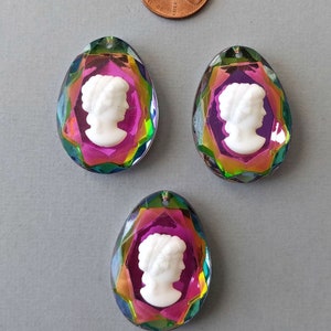 May include: Three iridescent teardrop-shaped pendants with a white cameo profile of a woman. The pendants have a faceted, multi-colored border and a small hole at the top for stringing. A penny is in the background.