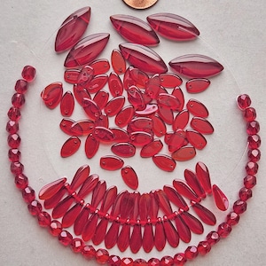 May include: A collection of red glass beads in various shapes and sizes. The beads include oval, teardrop, and faceted round shapes. Some beads are strung together, while others are loose. A penny is in the background.