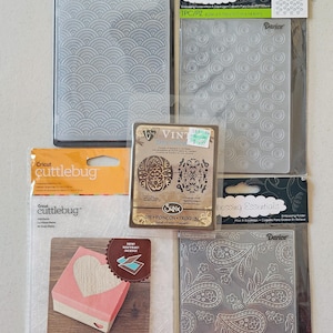 May include: A collection of embossing folders and die-cutting tools. The folders feature various patterns, including waves, circles, and paisley designs. One package includes a Cricut Cuttlebug die-cutting machine. The items are packaged in clear plastic.