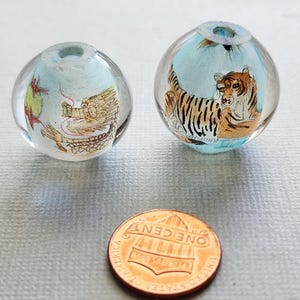 May include: Two clear glass beads with internal painted designs. One bead features a dragon, the other a tiger. The beads are near a penny for size comparison. The designs are set against a light blue background.