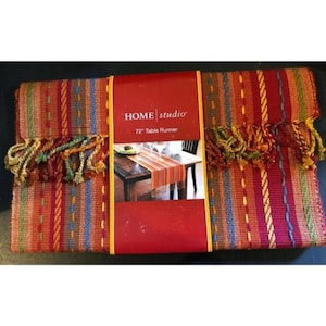 May include: A Home Studio 72-inch table runner. The runner is a mix of red, orange, green, and blue vertical stripes. The ends have decorative tassels in matching colors. The packaging is red with the Home Studio logo.