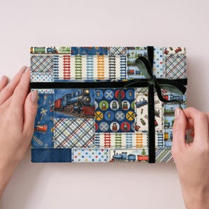 K&ouml;nnte beinhalten: Eine rechteckige Geschenkbox, die mit Patchwork-Stoff mit Illustrationen von Z&uuml;gen, Bahn&uuml;berg&auml;ngen und Gleisdesigns umwickelt ist. Die Box ist mit einem schwarzen Samtband und einer dunkelgr&uuml;nen Schleife gesichert.