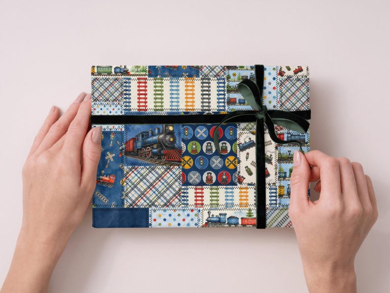 K&ouml;nnte beinhalten: Eine rechteckige Geschenkbox, die mit Patchwork-Stoff mit Illustrationen von Z&uuml;gen, Bahn&uuml;berg&auml;ngen und Gleisdesigns umwickelt ist. Die Box ist mit einem schwarzen Samtband und einer dunkelgr&uuml;nen Schleife gesichert.