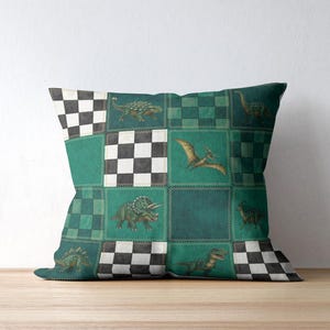 May include: Square throw pillow with a patchwork design featuring dinosaurs in shades of green, teal, and black. The pillow has a checkerboard pattern and various dinosaur illustrations, including a Triceratops and a Pterodactyl.
