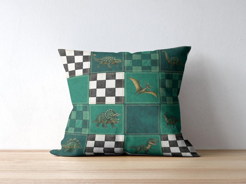 May include: Square throw pillow with a patchwork design featuring dinosaurs in shades of green, teal, and black. The pillow has a checkerboard pattern and various dinosaur illustrations, including a Triceratops and a Pterodactyl.