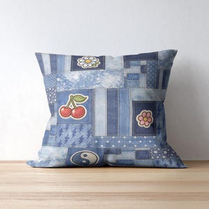 Op de afbeelding: Vierkantig sierkussen met een patchwork denim design. Het kussen heeft verschillende denim patches in blauwtinten, versierd met geborduurde kersen, madeliefjes, bloemen en yin-yang ontwerpen. Het kussen ligt op een houten oppervlak.
