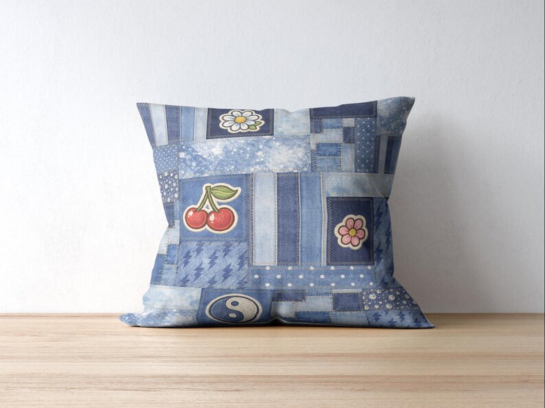 Op de afbeelding: Vierkantig sierkussen met een patchwork denim design. Het kussen heeft verschillende denim patches in blauwtinten, versierd met geborduurde kersen, madeliefjes, bloemen en yin-yang ontwerpen. Het kussen ligt op een houten oppervlak.