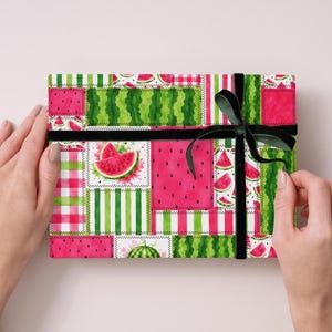 May include: A rectangular gift box wrapped in paper featuring a patchwork design of watermelon slices, seeds, and green stripes. The box is tied with a black velvet ribbon tied in a bow on the right side, showcasing a vibrant summer fruit theme.