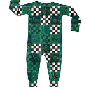 May include: Green and black dinosaur-themed one-piece pajama set. The pajamas feature a checkered pattern with various dinosaur illustrations. The long-sleeved, full-length sleepwear is designed for comfort and warmth. The color palette includes shades of green, black, and white.