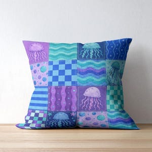 May include: Square cushion with a patchwork design featuring jellyfish, waves, and geometric patterns in shades of blue, purple, and teal. The cushion is placed on a wooden surface.