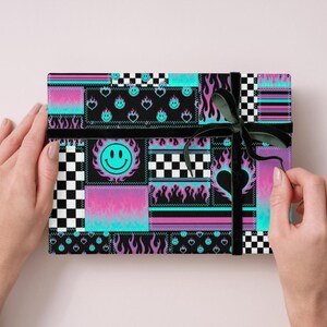 K&ouml;nnte beinhalten: Eine rechteckige Geschenkbox, eingewickelt in Papier mit einem Patchwork-Design aus schwarz-wei&szlig;em Schachbrettmuster, neonblauen Flammen, violetten Farbverl&auml;ufen und t&uuml;rkisfarbenen Smileys, gebunden mit einem d&uuml;nnen schwarzen Band.