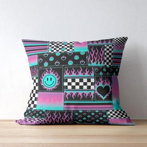 K&ouml;nnte beinhalten: Ein quadratisches Dekokissen mit einem Patchwork-Design in Schwarz, Pink und Cyanblau. Das Muster besteht aus Schachbrettmustern, Flammenmotiven, Smileys und Herzformen, die in einem Raster mit angedeuteten N&auml;hten angeordnet sind.