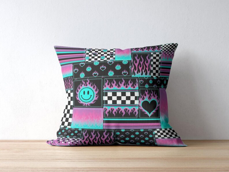 K&ouml;nnte beinhalten: Ein quadratisches Dekokissen mit einem Patchwork-Design in Schwarz, Pink und Cyanblau. Das Muster besteht aus Schachbrettmustern, Flammenmotiven, Smileys und Herzformen, die in einem Raster mit angedeuteten N&auml;hten angeordnet sind.