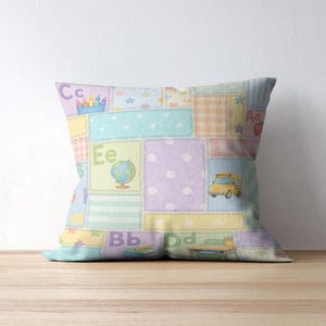 May include: A square cushion with a patchwork design in pastel colours. The cushion features squares with letters, a school bus, a globe, and pencils. The cushion is on a wooden surface.
