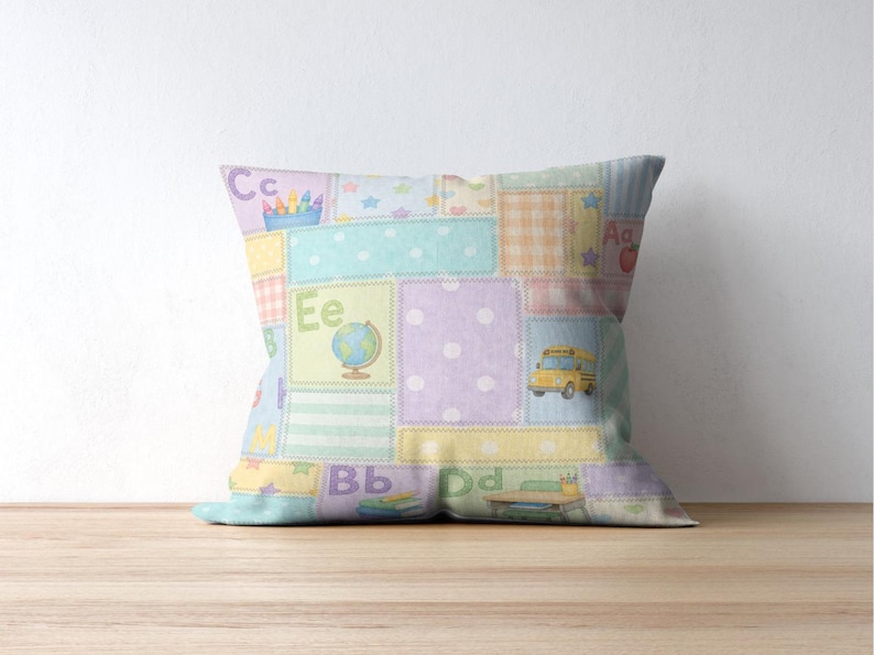 May include: A square cushion with a patchwork design in pastel colours. The cushion features squares with letters, a school bus, a globe, and pencils. The cushion is on a wooden surface.
