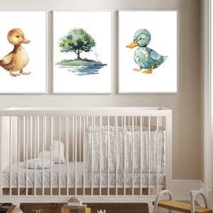 May include: Three watercolor prints featuring a brown duck, a blue duck, and a green tree with a blue pond. The prints are framed in white and hung on a white wall above a white crib.